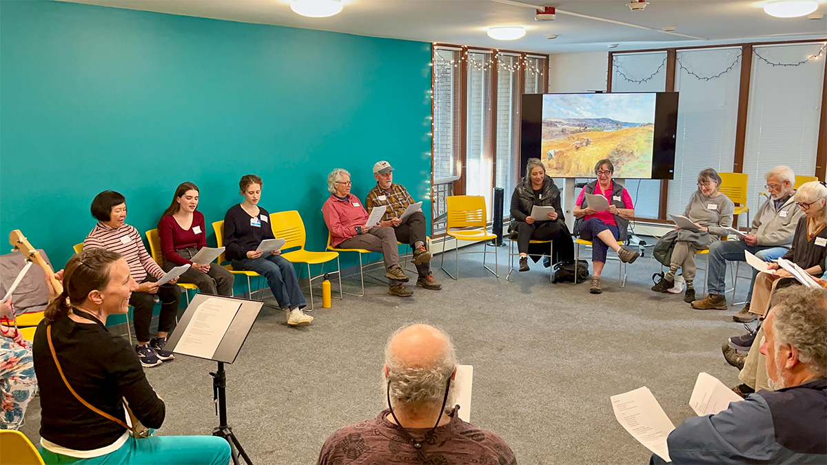 A group of about 15 people seated in a circle on chairs in a medium sized room. They are holding papers and following along as one person plays a guitar.