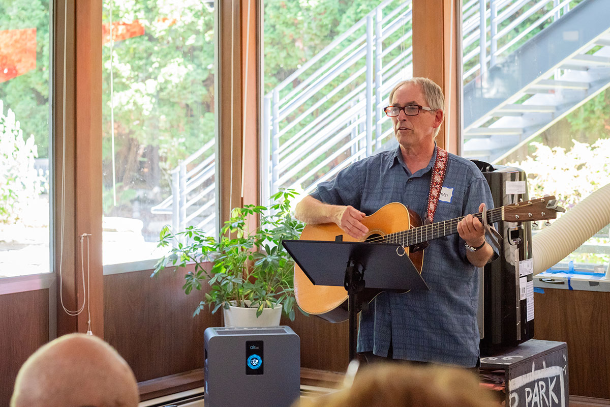 A person standing and playing an acoustic guitar for a small seated audience in a room with large windows