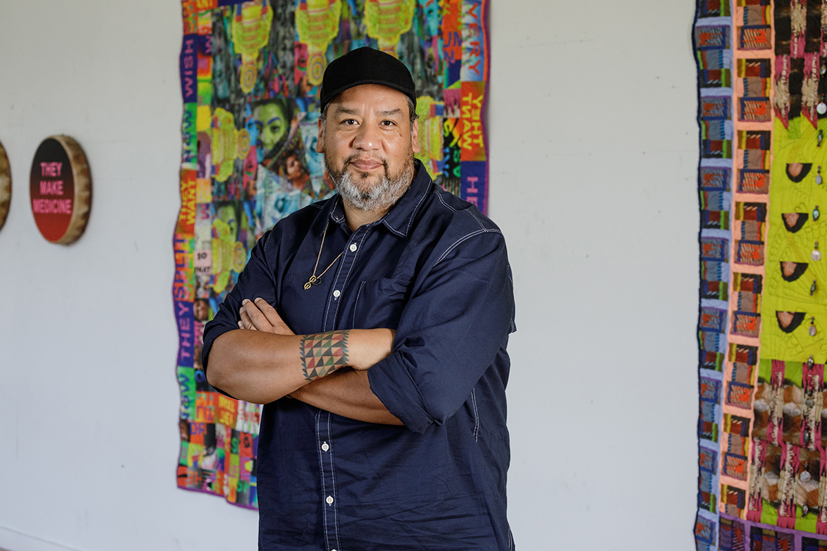 A person standing with their arms crossed in front of hanging textile artworks on a wall