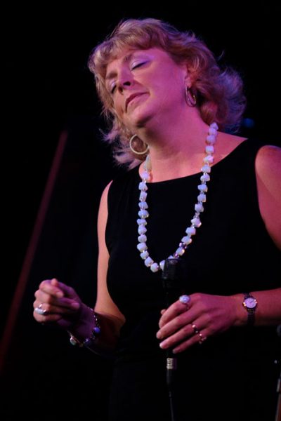 Photo of Greta Matassa, a woman with curly blonde hair wearing a black dress and white necklace, holding a microphone in a spotlight