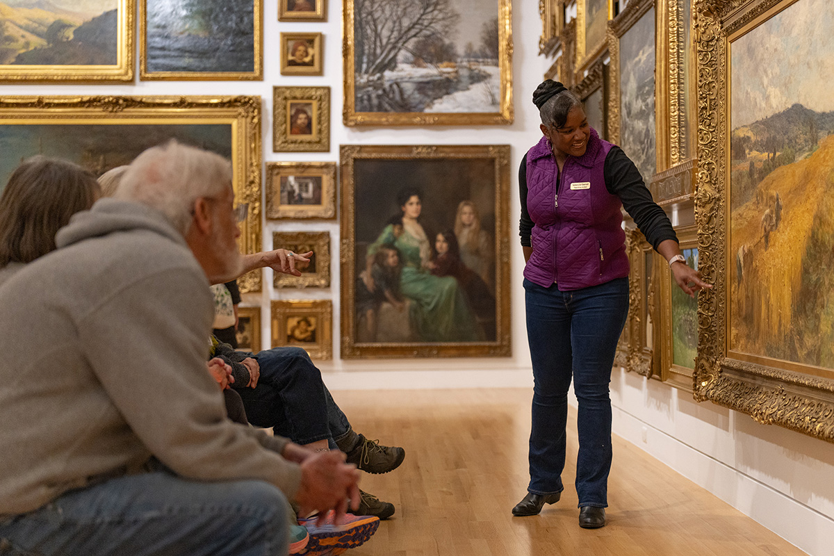 A woman wearing a purple vest pointing at a painting in a room filled with paintings while leading a discussion. Older adults are seating and watching, one is pointing towards the painting as well.