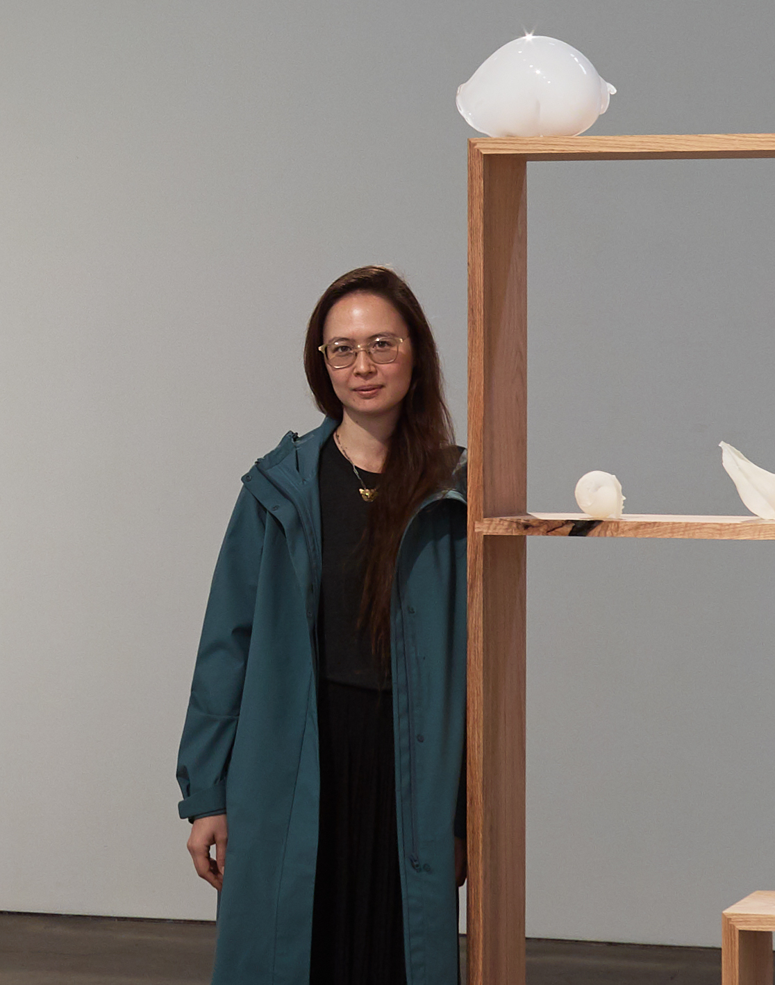 Woman with glasses and long dark hair wearing a blue/green jacket and black dress leaning against a wooden shelf displaying glass sculptures