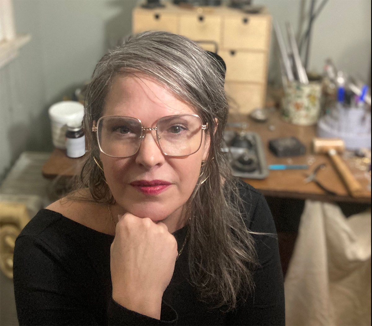 A woman wearing large glasses in a workshop or studio looking at the viewer and resting her chin on her hand