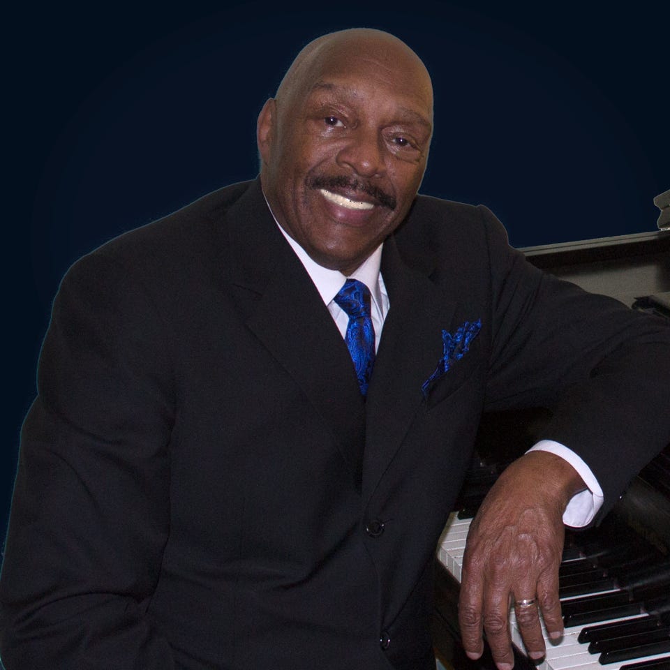 A formal portrait style photo of a black man wearing a suit and blue tie leaning against a piano