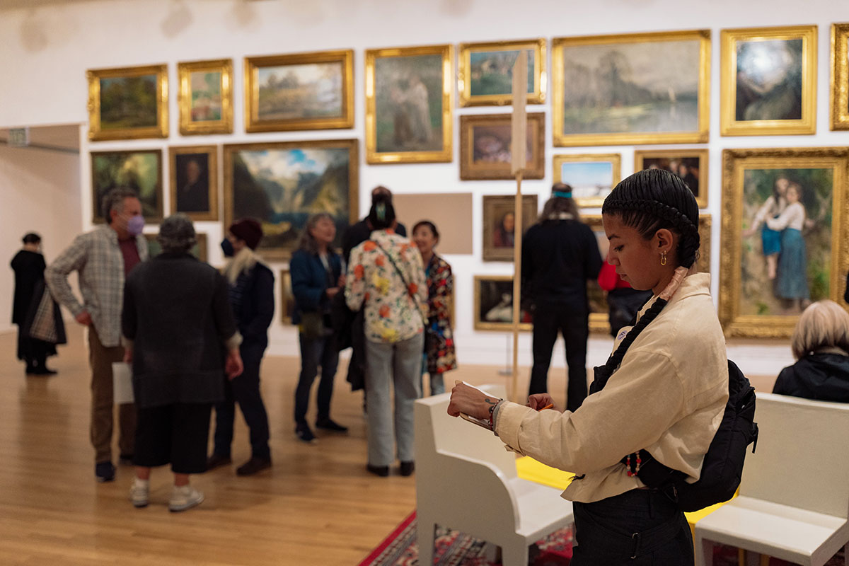 A woman wearing a white shirt and a black bag is writing on a paper on a clipboard while standing in front of a wall of framed landscape paintings in an art gallery. Several other visitors are gathered around, observing the artwork. The gallery floor is wood, and the room is bathed in soft light.