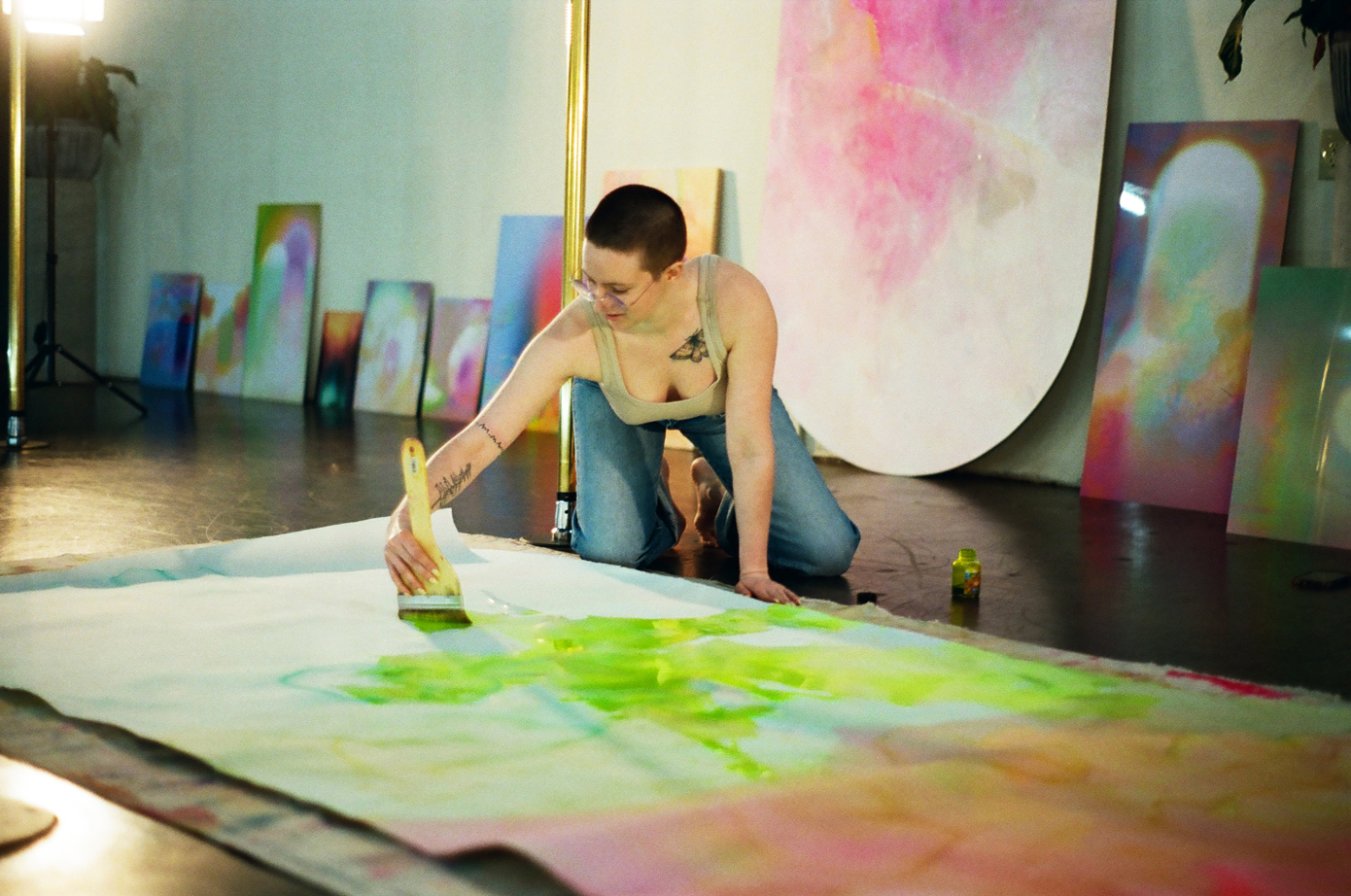 Photograph of Lydia Jewel Gerard, a person with short dark hair, kneeling next to an artwork on which she is painting with a large brush