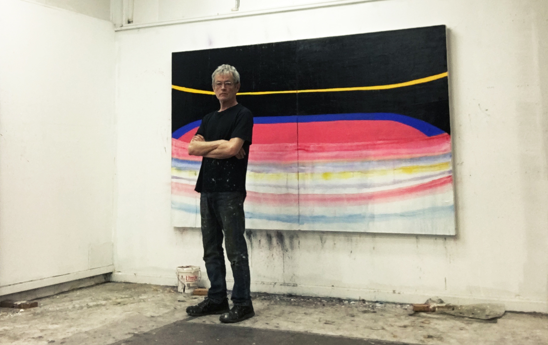 Photo of Ken Kelly, a man with short gray hair and glasses, looking at the camera, standing in front of a painting of his in his studio