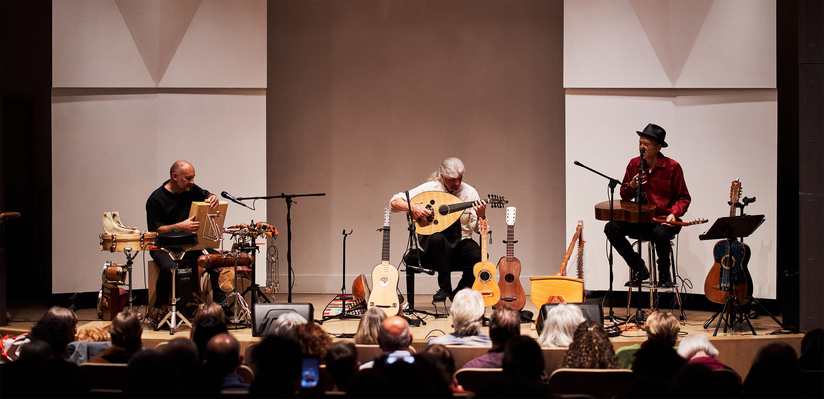 Three people performing music with older looking, mostly stringed instruments on a stage in front of a seated audience