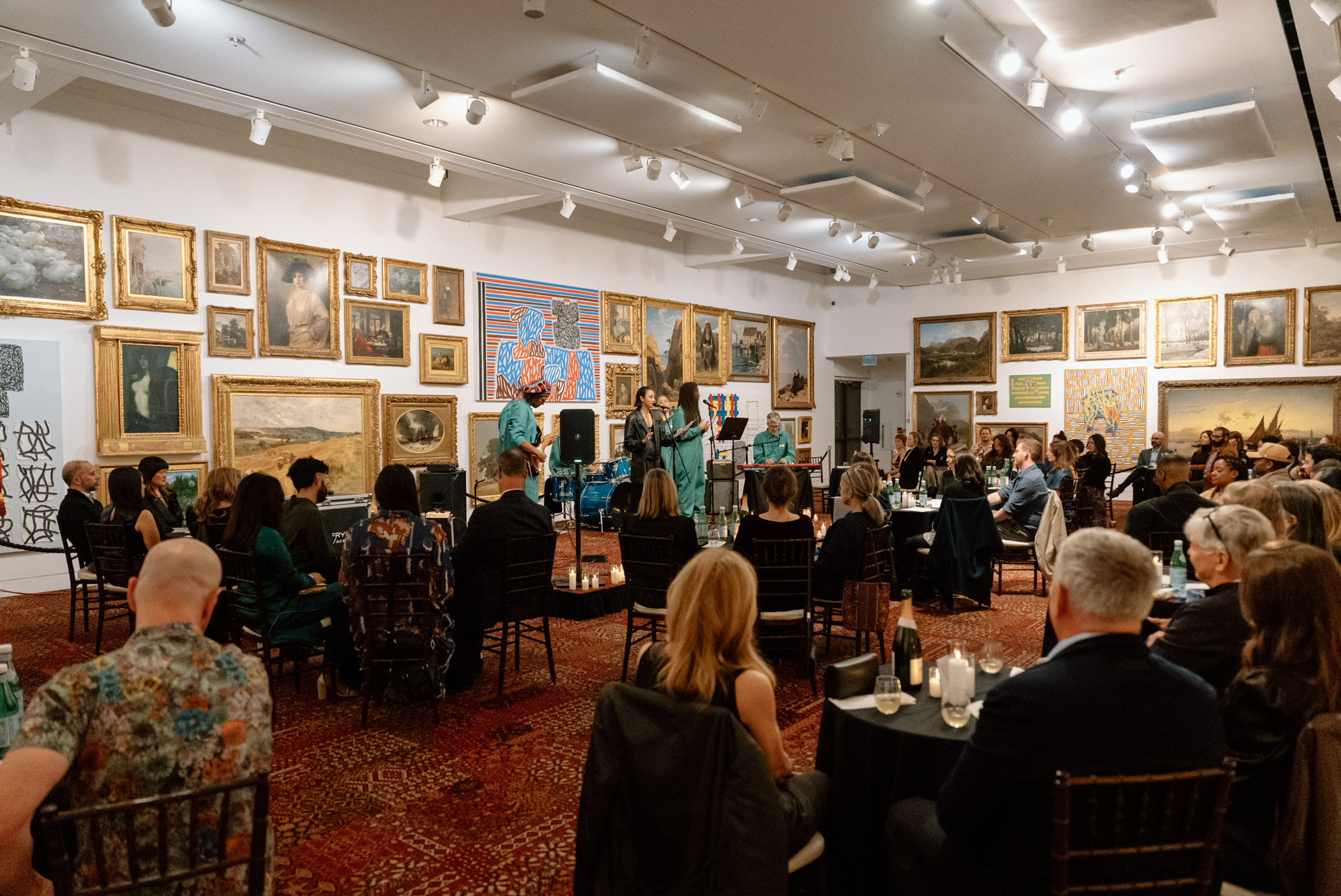 A band performing on a low stage for an audience seated at small tables in a large gallery space