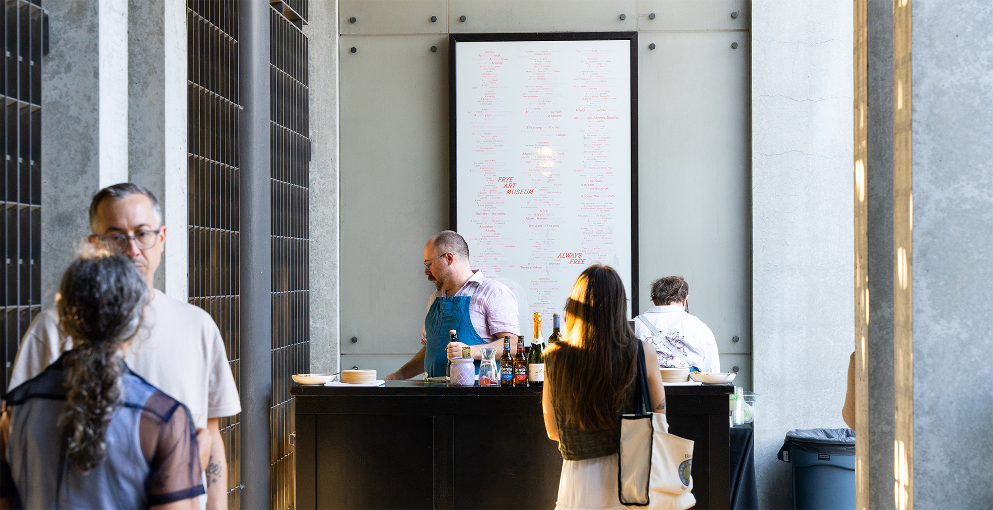 People standing in a courtyard with slanted evening sun near a temporary bar setup. A poster with a text design is on the wall behind the bar.