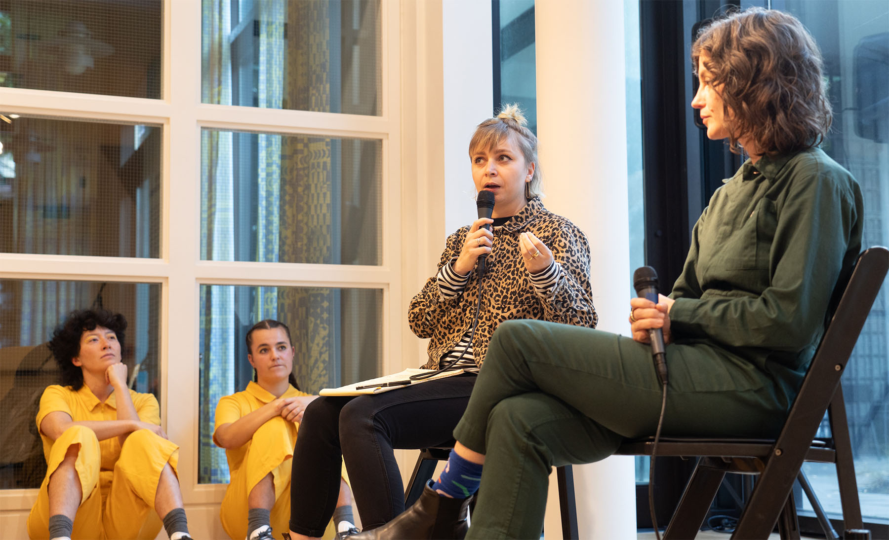 Two seated women talking with microphones to an out of frame audience, while a pair of people in yellow jumpsuits sit on the floor against a wall and watch them.