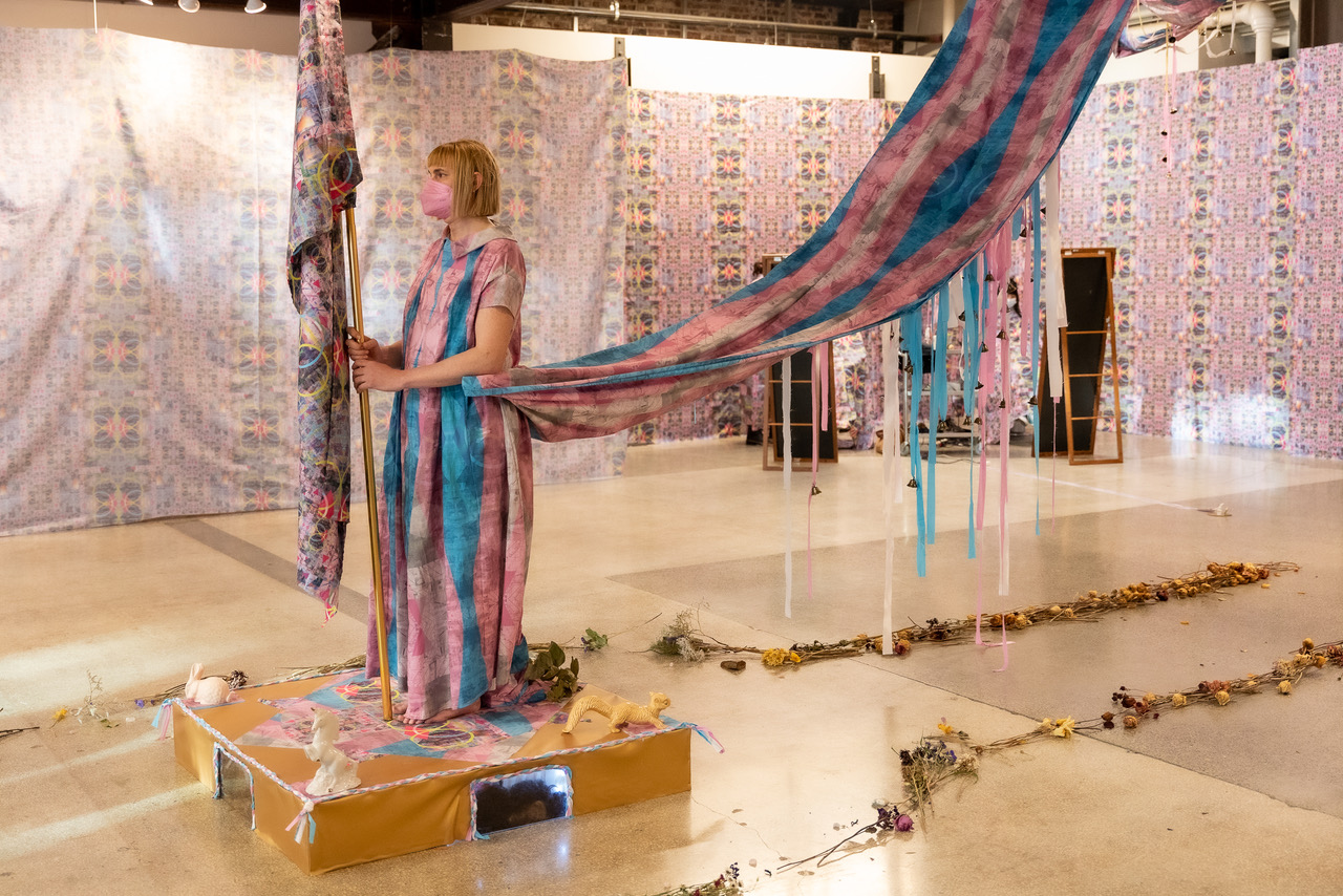 Photograph of Molly Jae Vaughan activating her installation "Safety in Numbers" which features many textiles, dried flowers, and found objects.