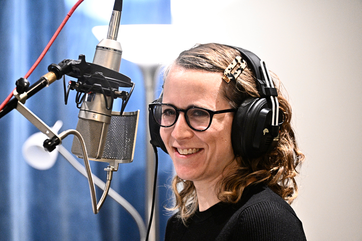Photo of Erin Langner, a woman with shoulder length brown hair and glasses, smiling in front of a microphone in a recording studio