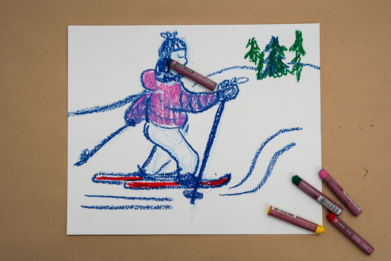 Photo of a piece of paper with a skier and trees drawn on it and pastels sitting on top of the lower right corner of the paper