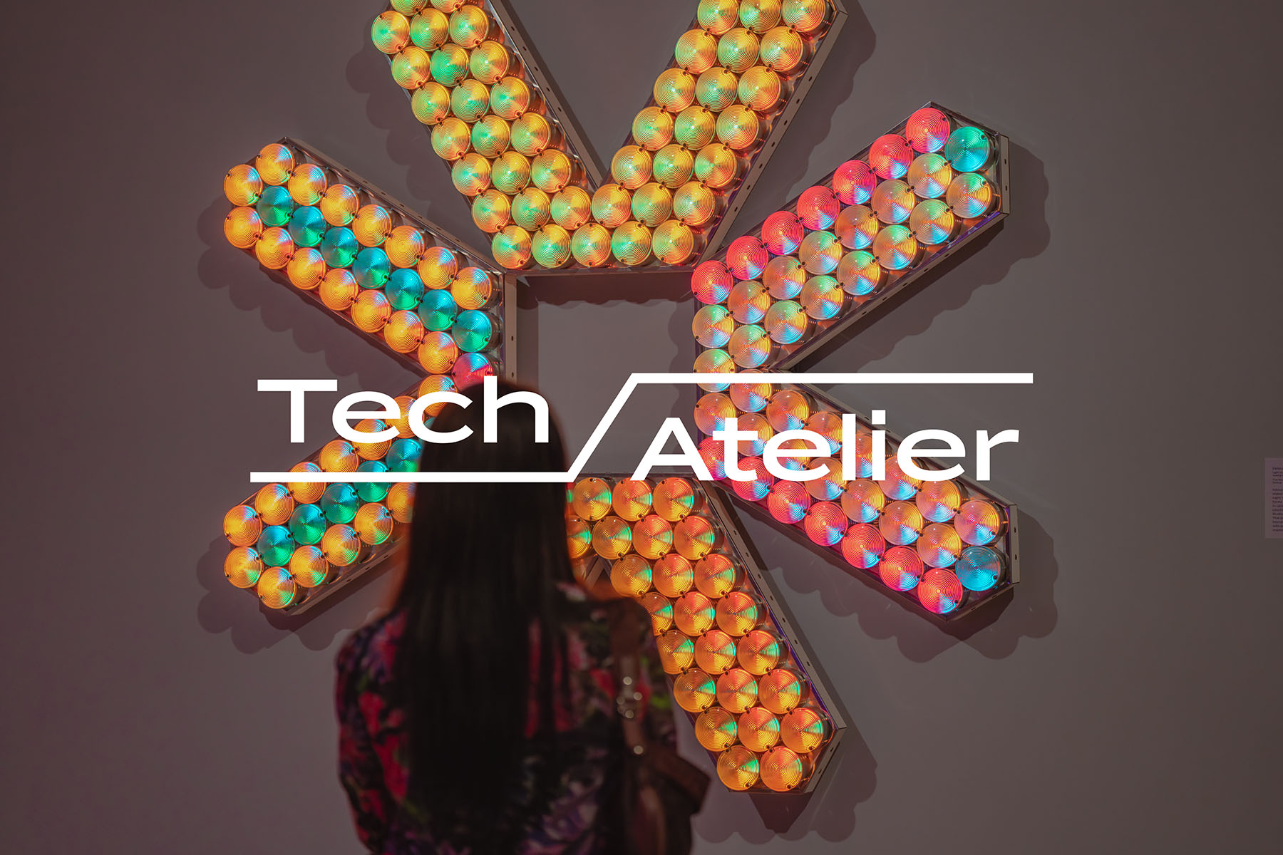 Text reading 'Tech Atelier' over a photo of a person looking at a geometric artwork with a number of colorful lightbulbs