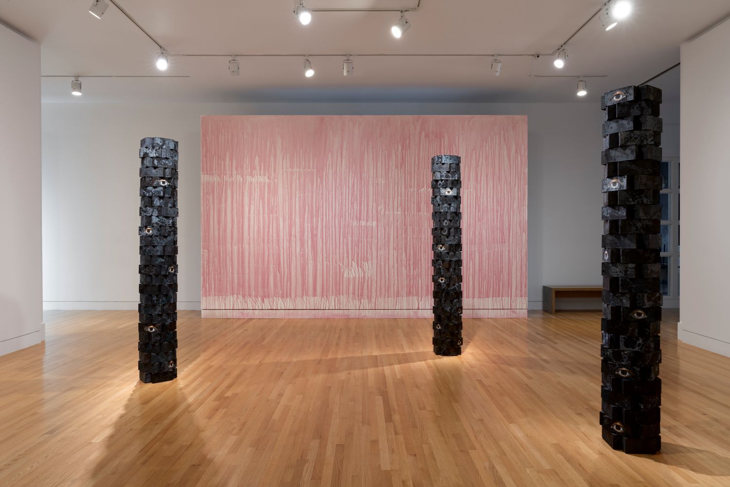 View of an art gallery with three sculptures made of bricks in the forefront and a wall painted with dripped beet juice in the background.