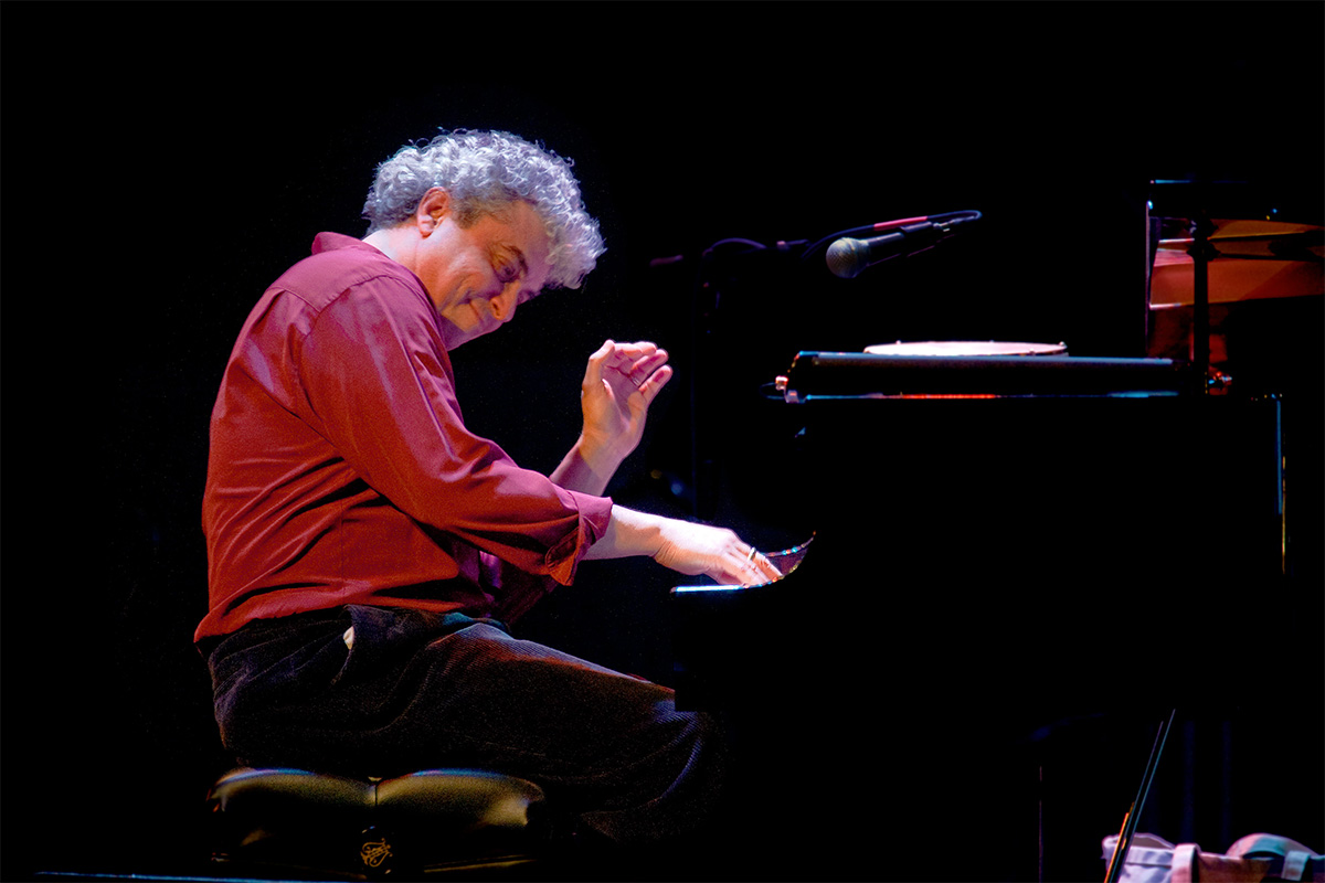 A man in a red shirt playing a grand piano in a large, dark room