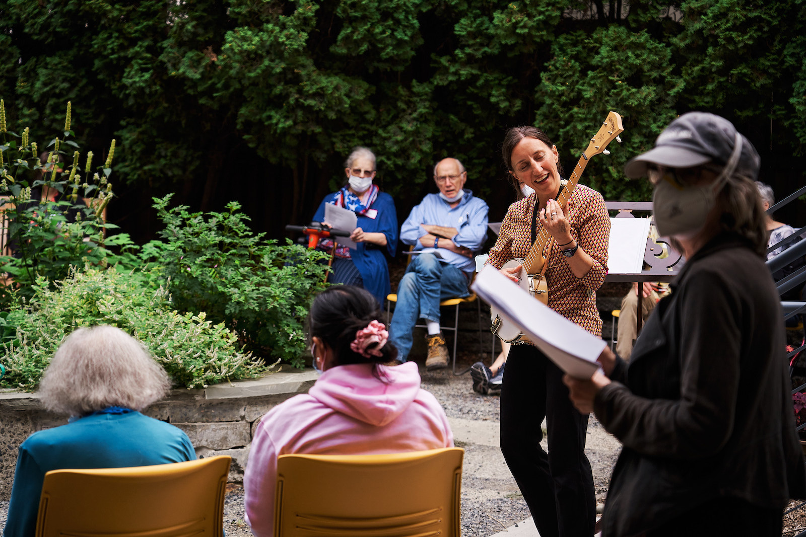 A person playing guitar standing in a garden, with older adults sitting on benches and seats around the area