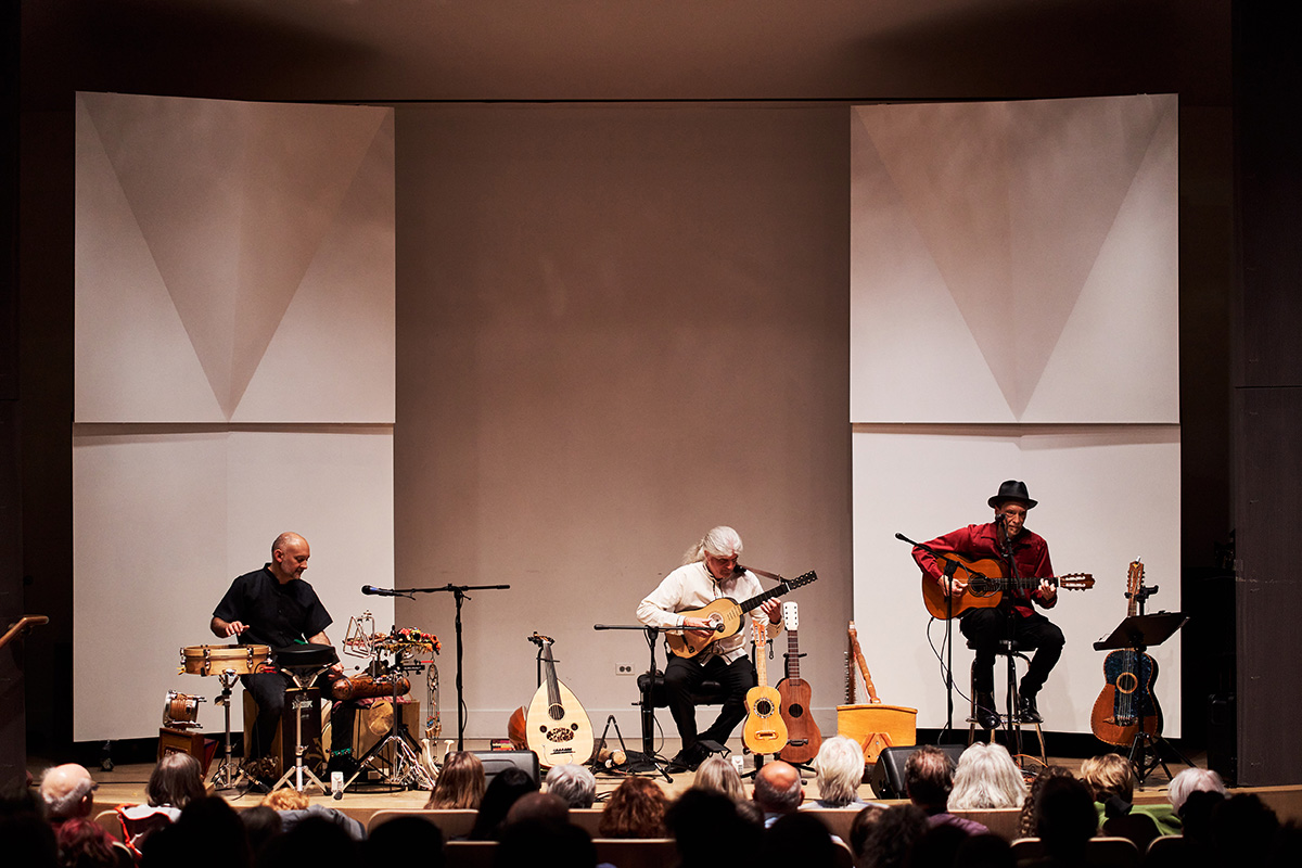 Three people performing music with older looking, mostly stringed instruments on a stage in front of a seated audience