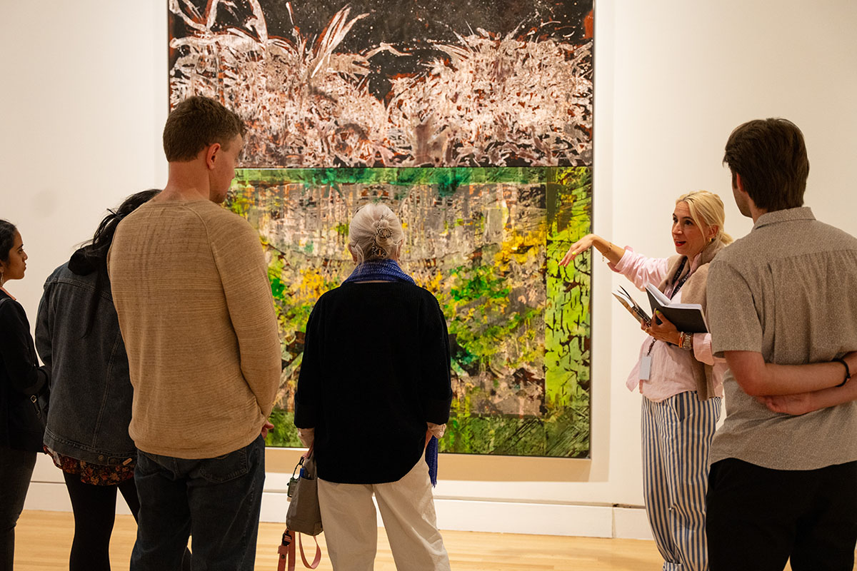 A woman standing next to a large abstract painting and speaking to a small group of people