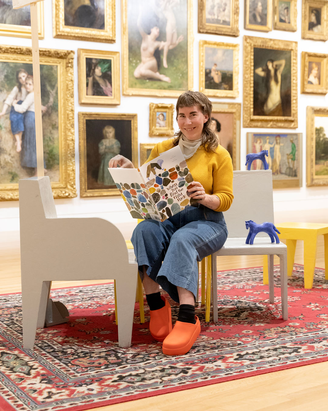 A person holding a large, open childrens book sits next to wooden, painted blue horse models on chairs in front of a wall of art