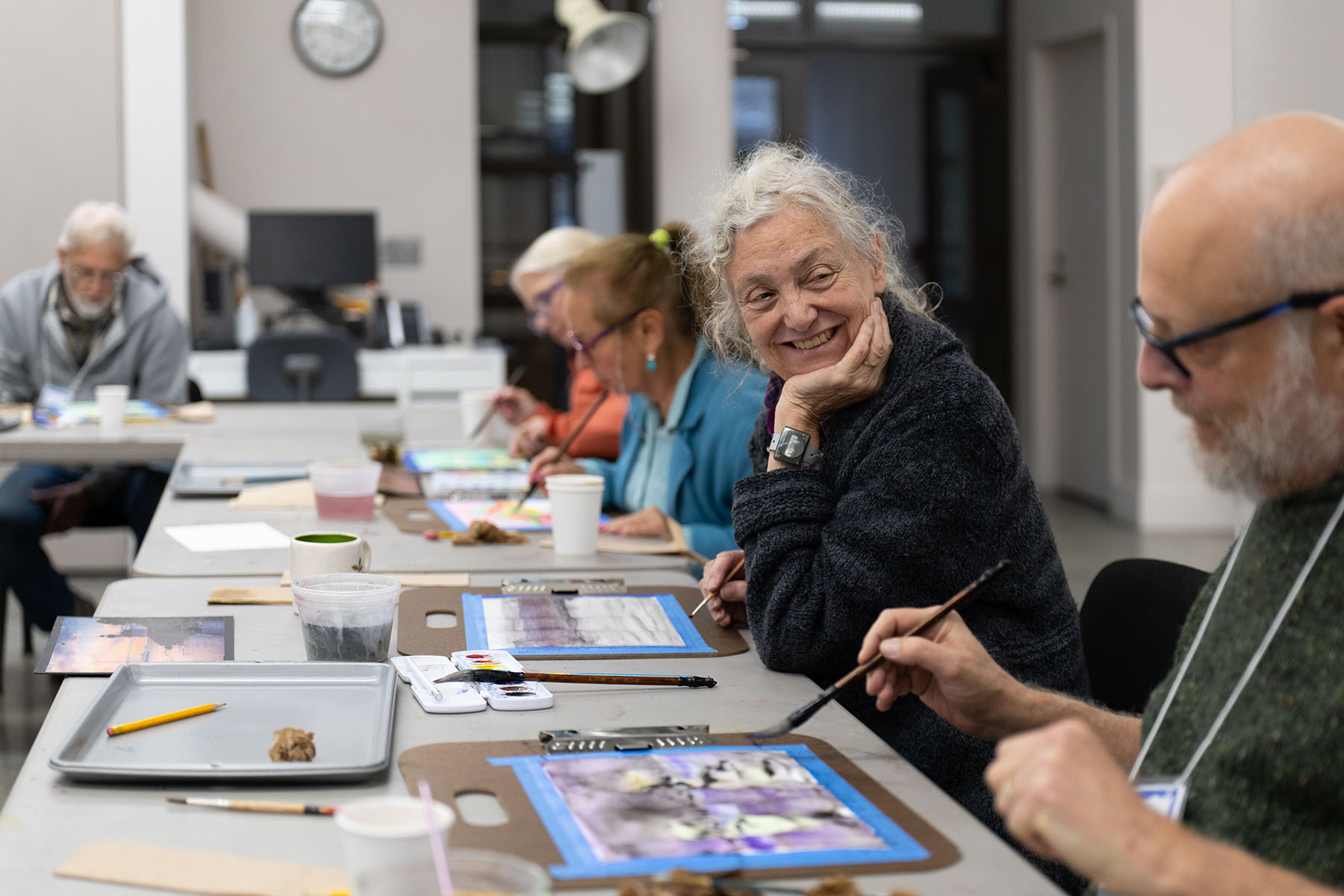 A group of older adults sitting at a long table working on an art project. A woman has her head turned and is similing at a person closer to the viewer