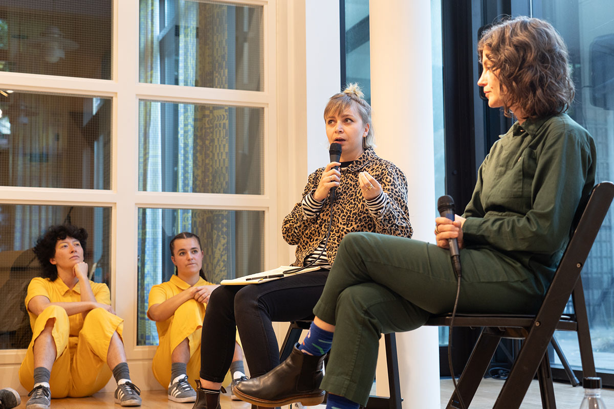 Two seated women talking with microphones to an out of frame audience, while a pair of people in yellow jumpsuits sit on the floor against a wall and watch them.