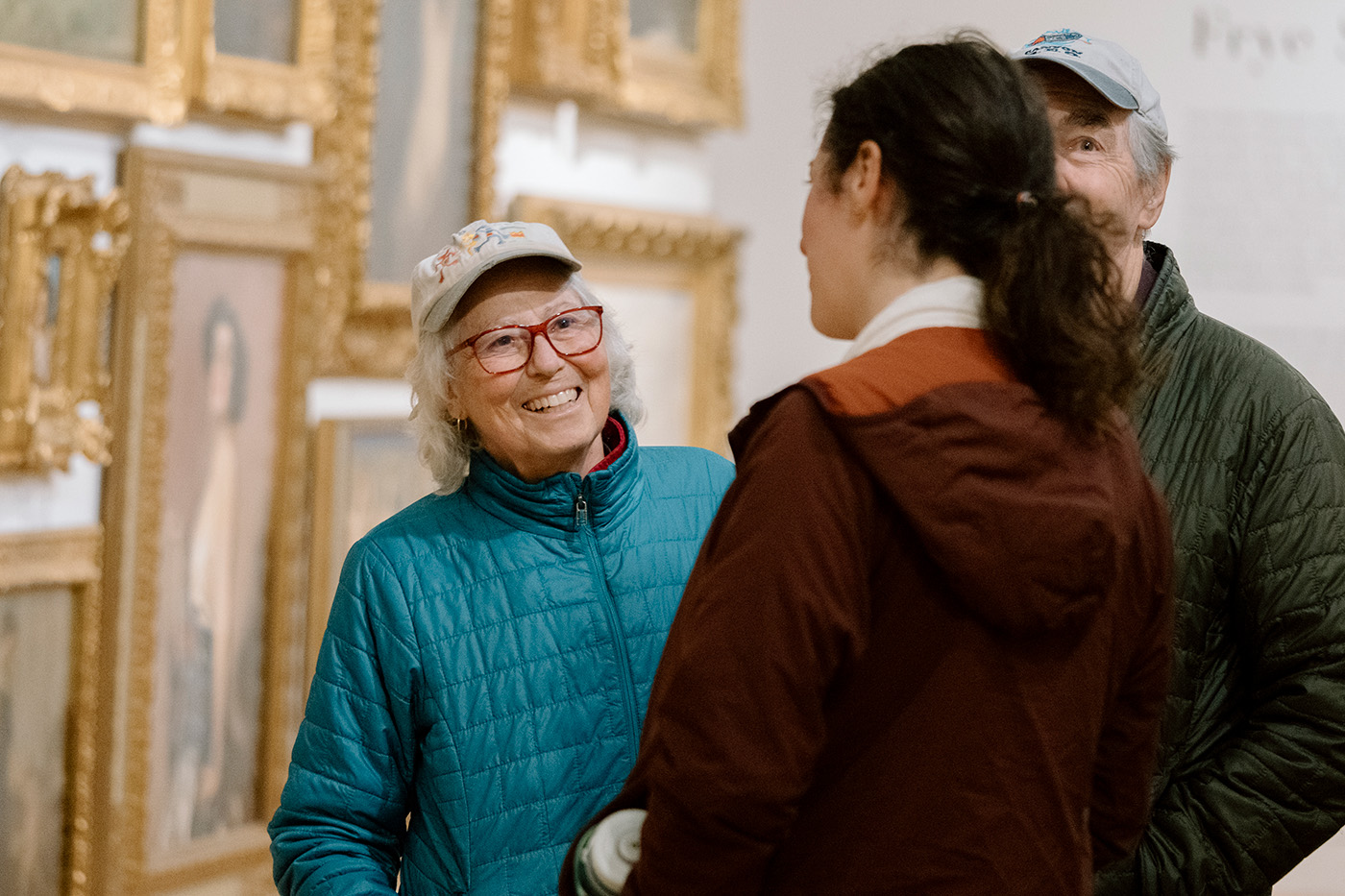 People standing an talking in front of a wall hung with many paintings in gold frames