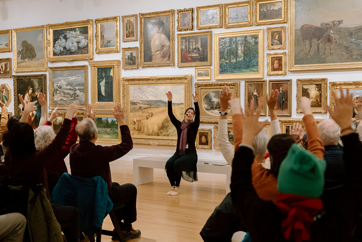 A person with their arms raised sits on a bench in front of a wall covered in paintings. There are many people seated and facing them.