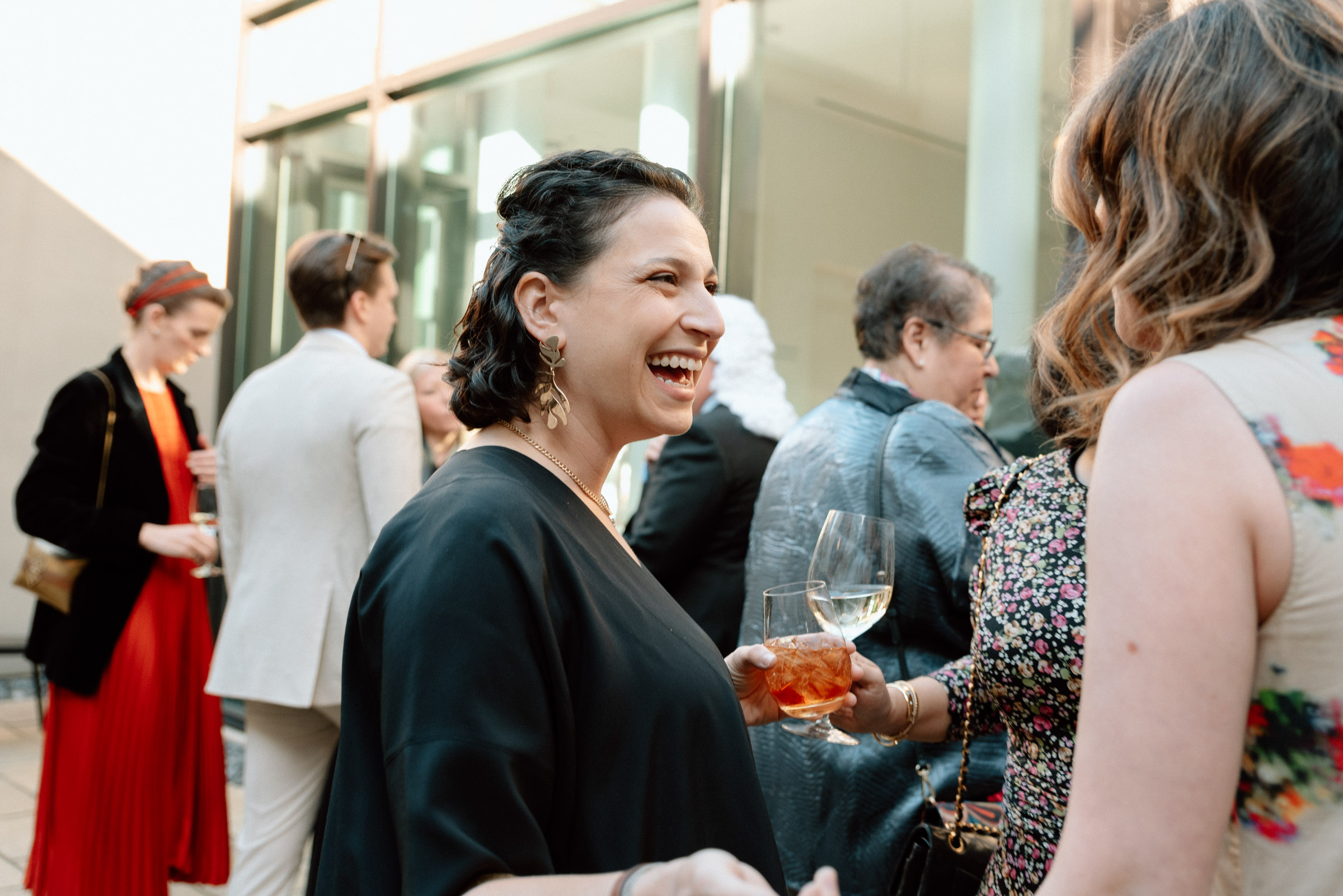 Well dressed people standing and chatting in a sunny courtyard. A woman in the center of the image is holding a cocktail