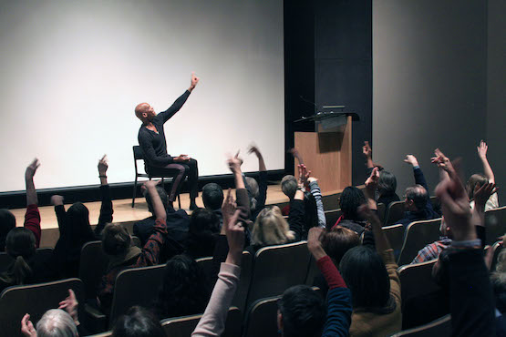 Photo of a group sitting in an auditorium with their hands raised in the air, and a presenter on stage