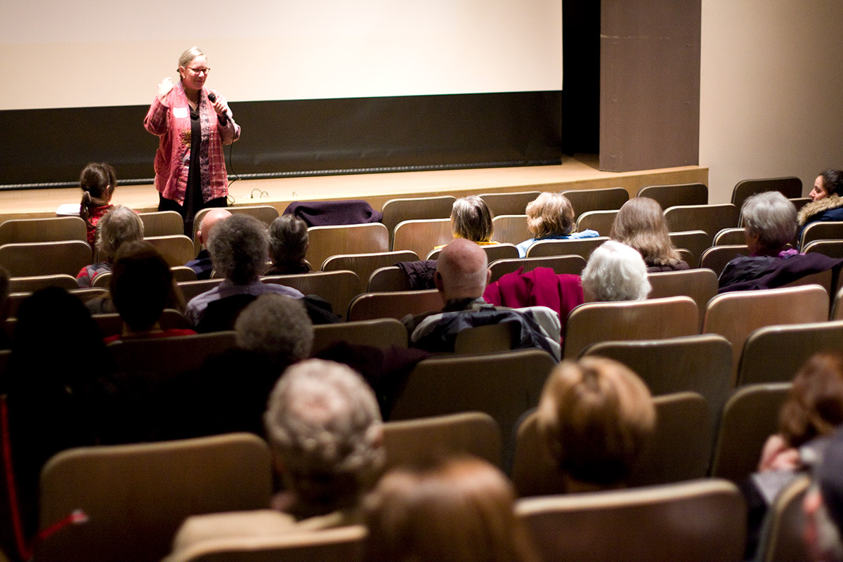 A woman speaking into a microphone in front of a seated audience in an indoor auditorium