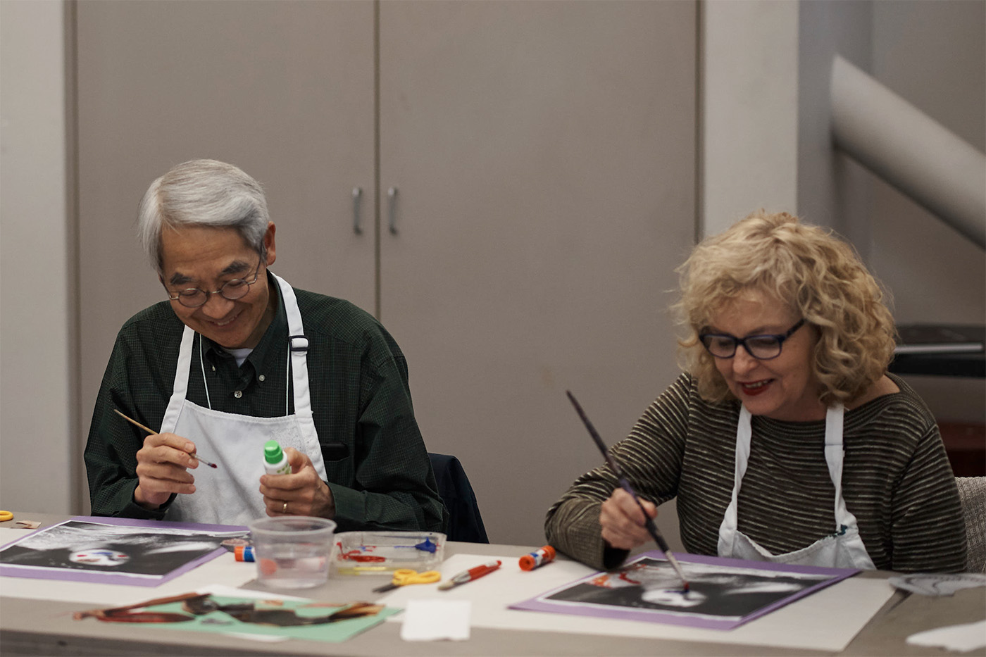 Two older adults sitting at a table wearing aprons and working on an art project