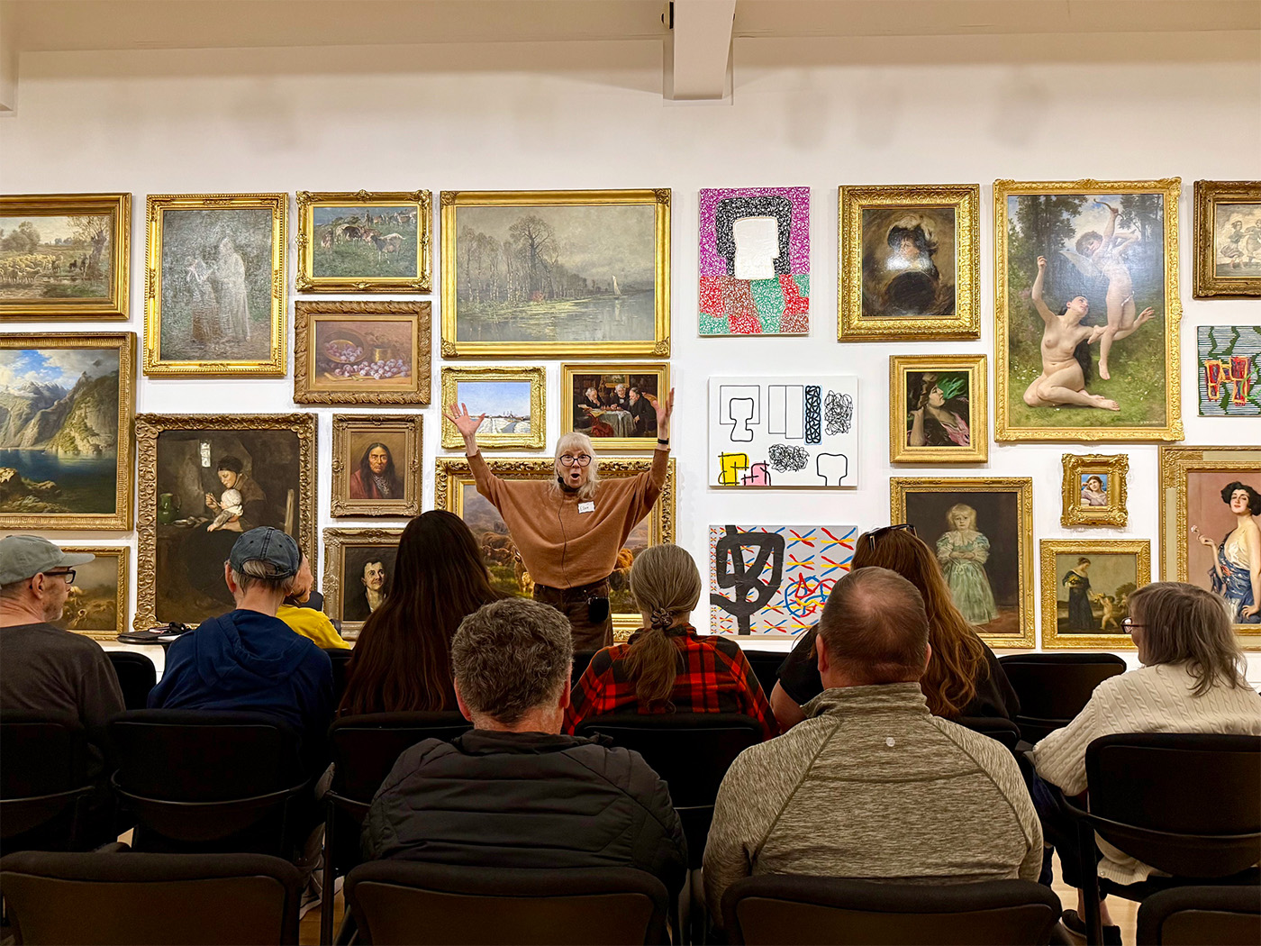 A woman gesturing broadly in front of a seated audience in a large art gallery space