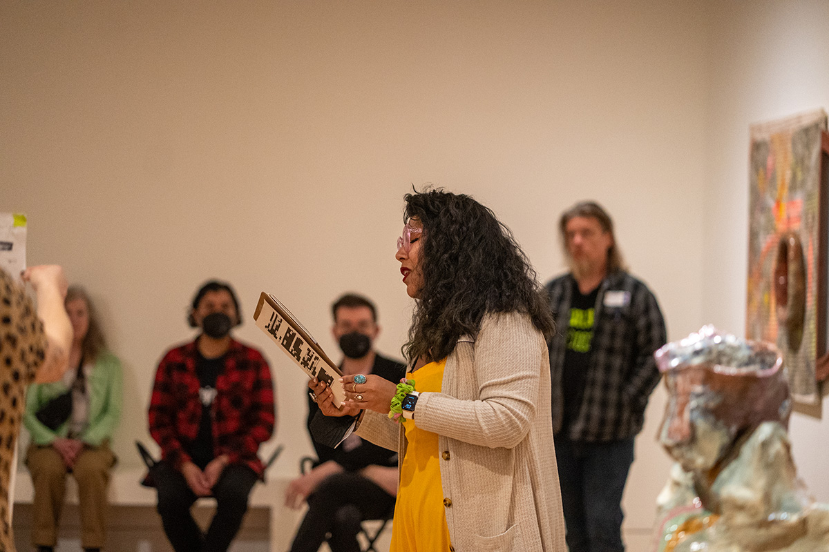 A woman in profile reading from a piece of paper in their hands to a group of people in an art gallery