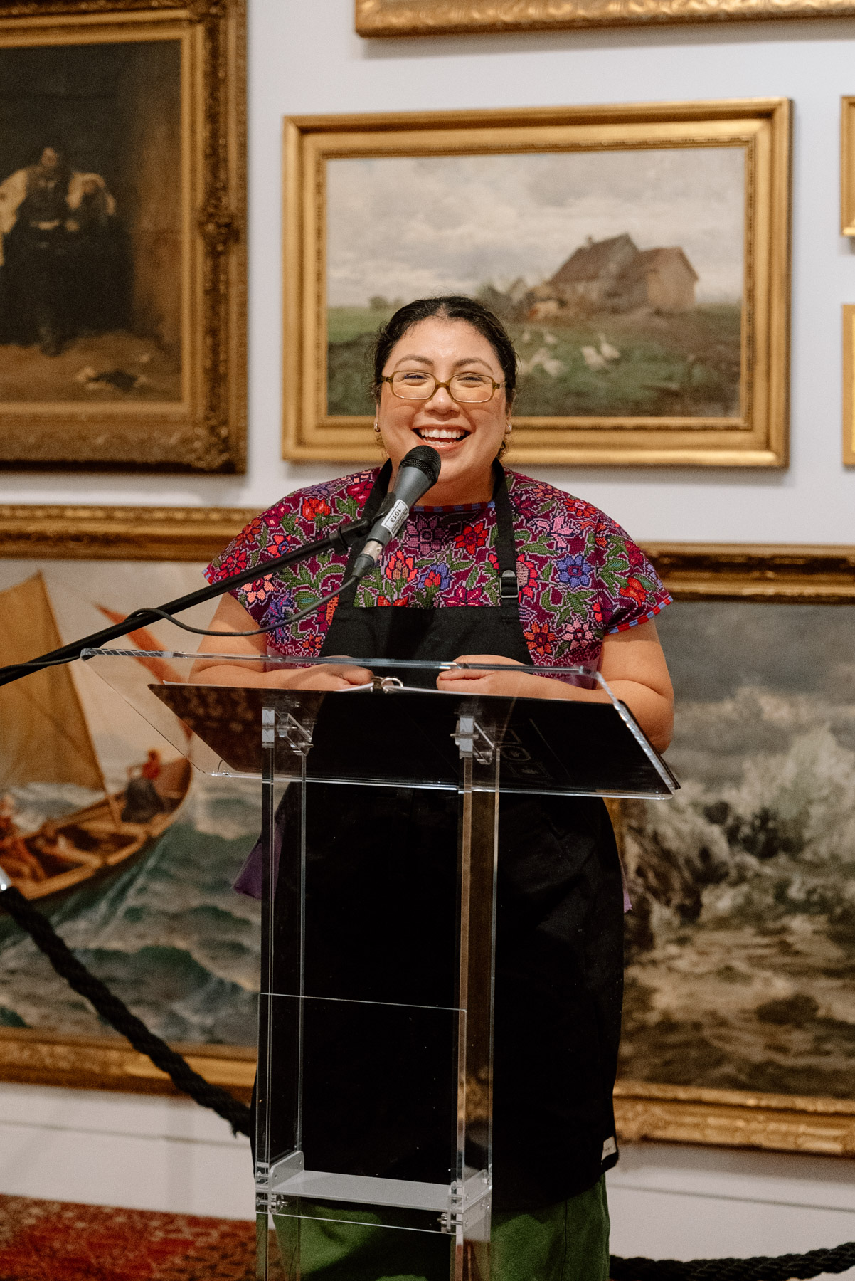 A woman smiling and talking into a mic at a lectern in front of oil paintings on a wall