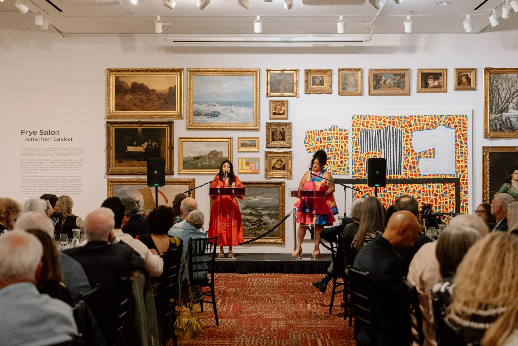 Two people on a low stage at lecterns talking to people seated at two long dinner tables in an art gallery space