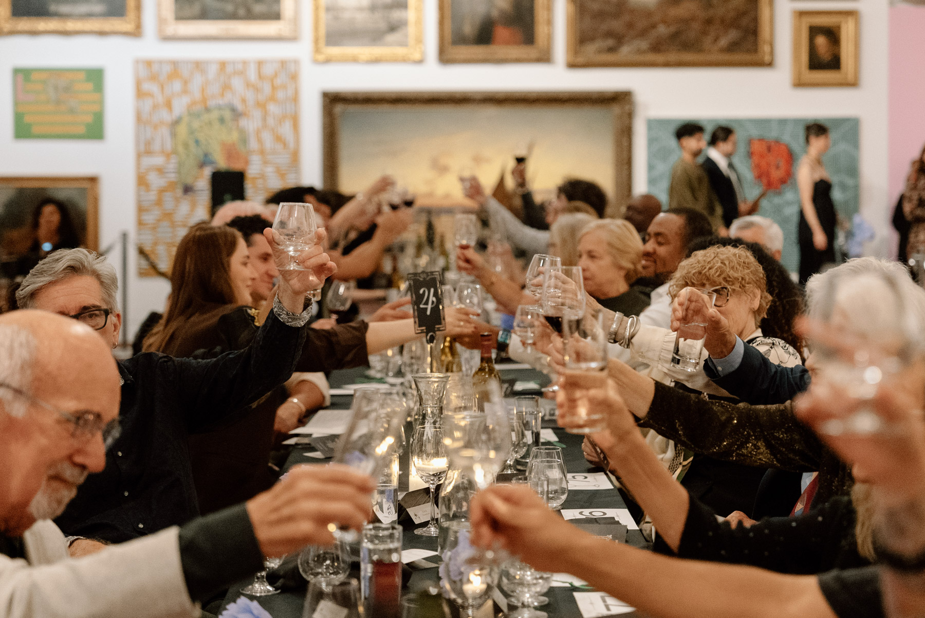 A large number of people seated at a long table in an art gallery space raising their glasses in a toast