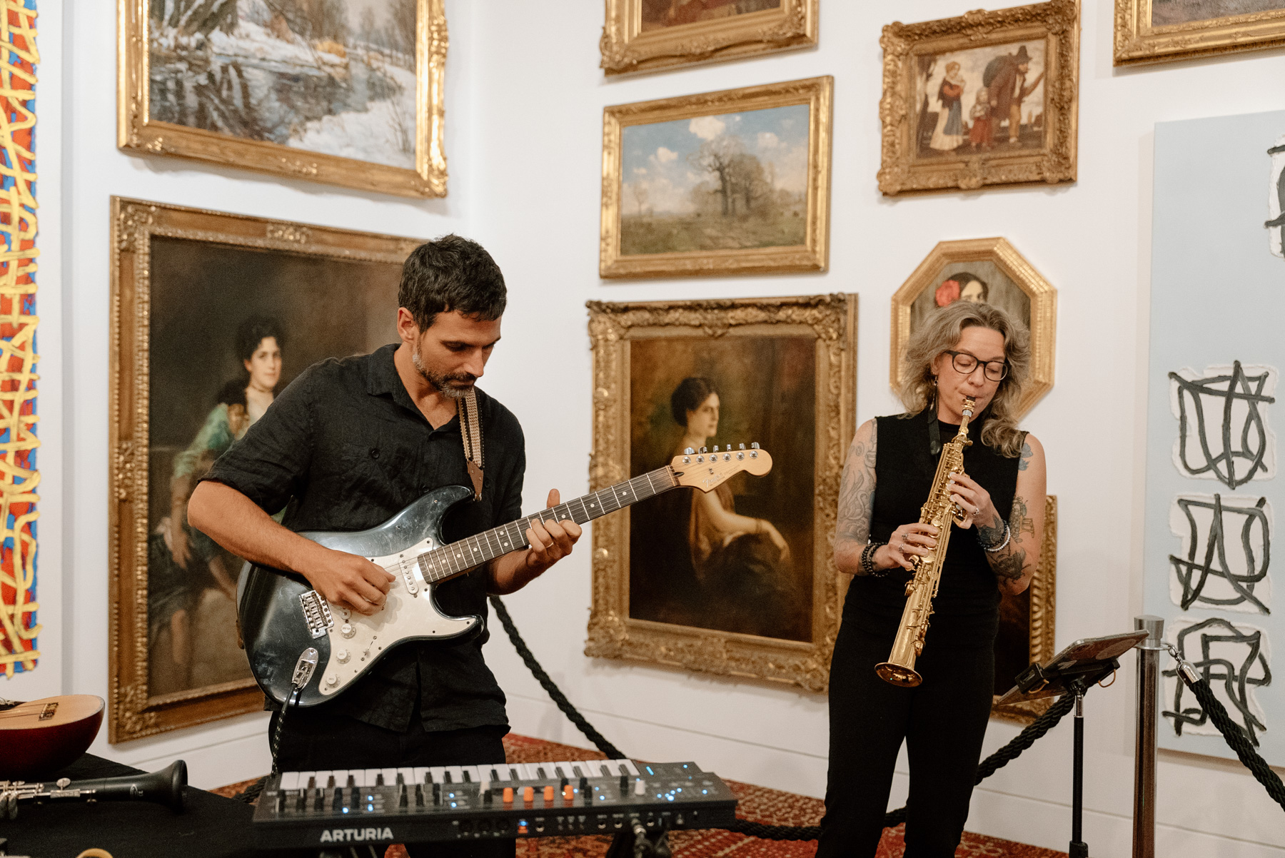 Two musicians playing in front of a corner of an art gallery covered in oil paintings in gold frames