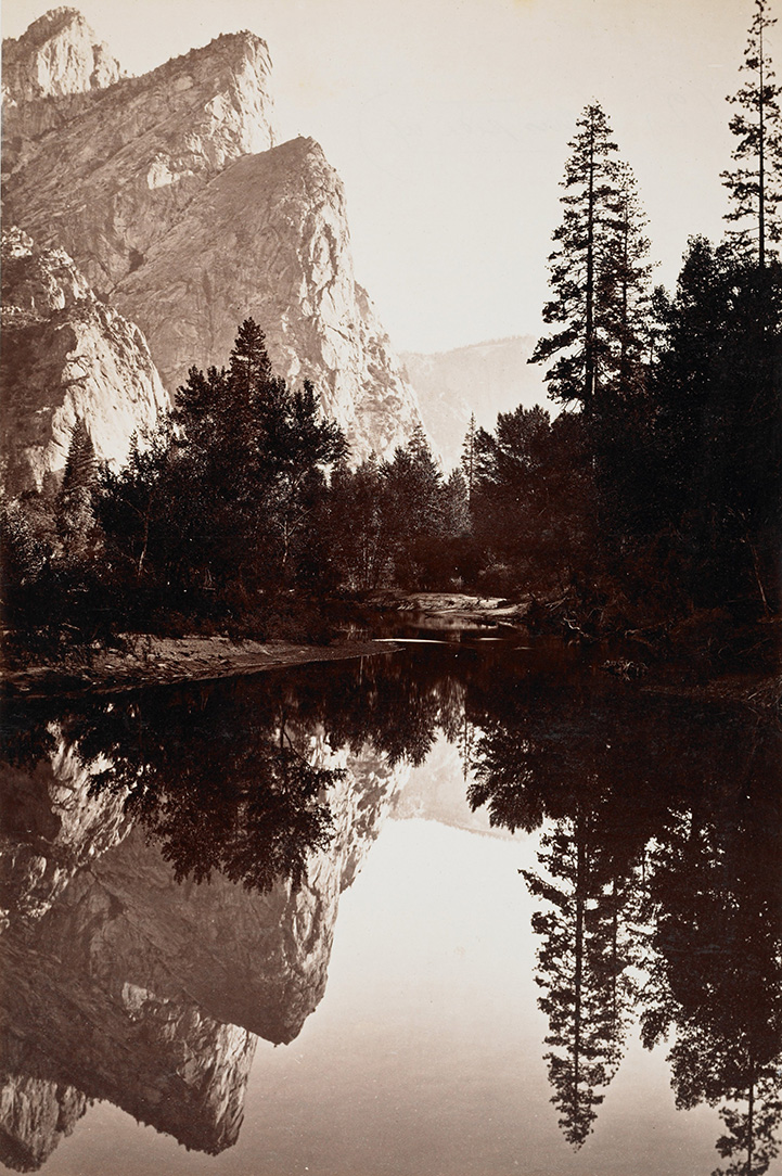 A sepia photo of a mountain lake surrounded by evergreen trees and jagged peaks