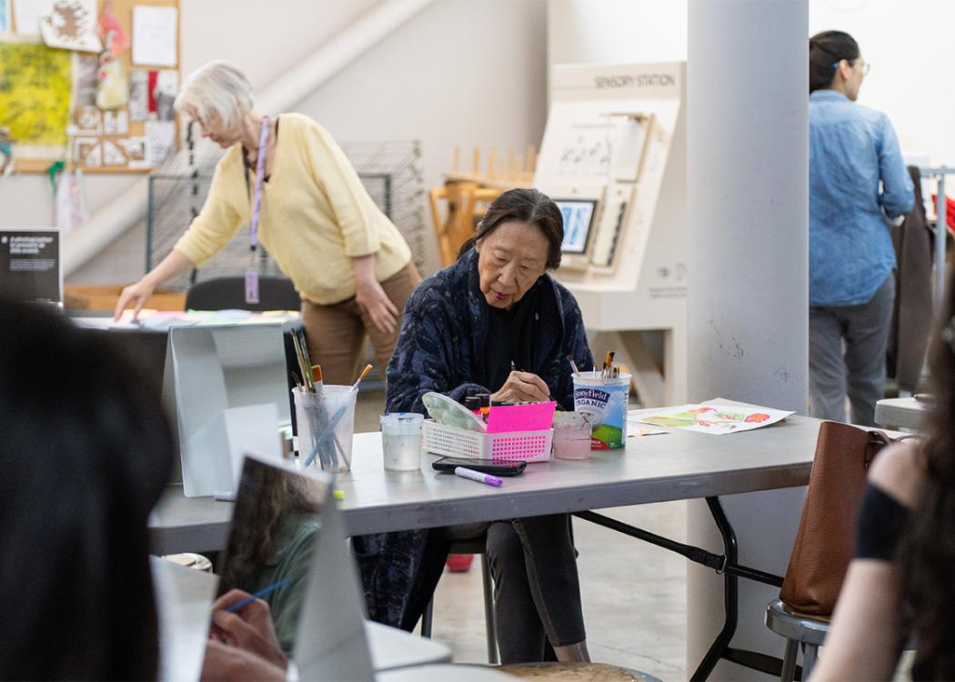 A person sitting and working on an art project. Other people are visible doing the same in other pages of the room.