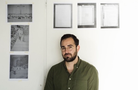 Man with dark hair and beard, dressed in a green button-down shirt, standing in his studio, surrounded by archival scans affixed to the wall