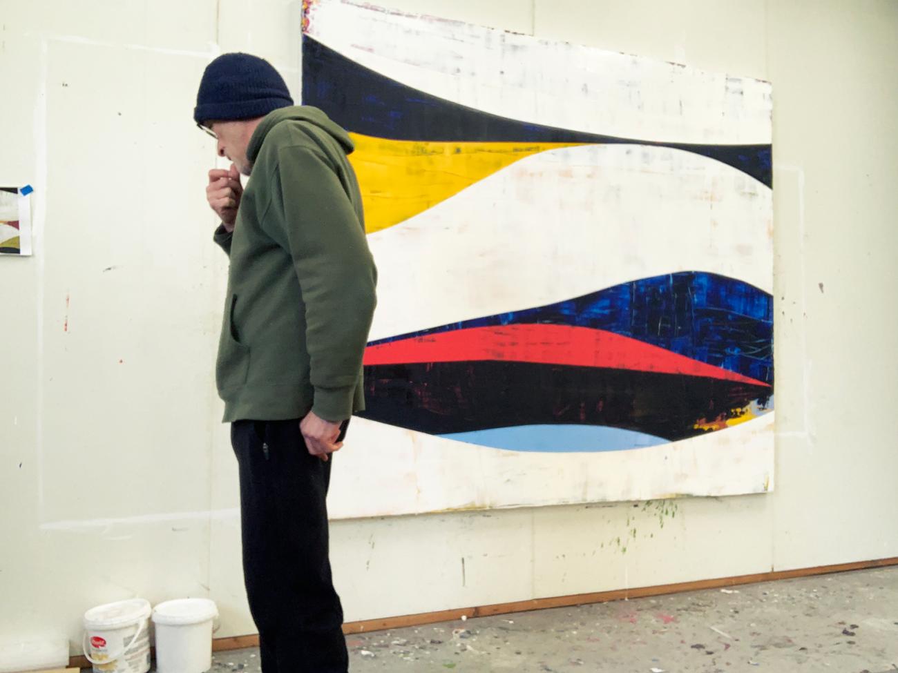 Photo of Ken Kelly, a man wearing a hoody and a beanie, working in his studio with one of his paintings on the wall behind him