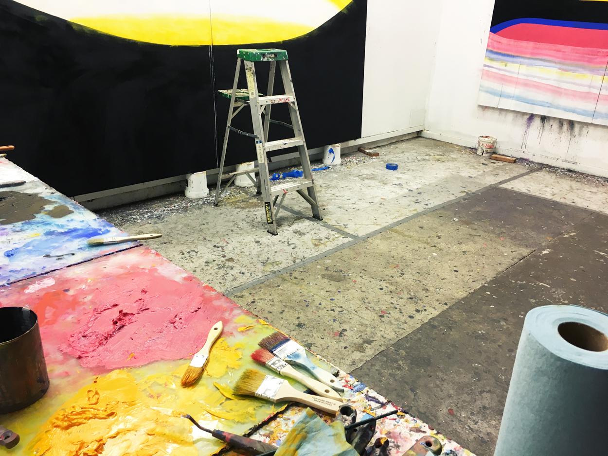 Photo of Ken Kelly's painting studio with large paintings on the walls and a table with paint and paintbrushes on it on the left side