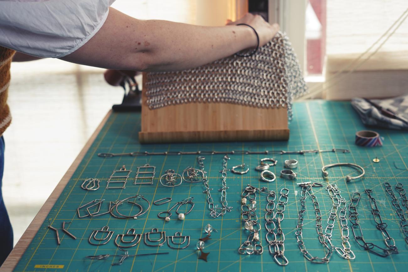 Photo of silver jewelry laid out on a workspace and a piece of chainmail is being laid over a stand