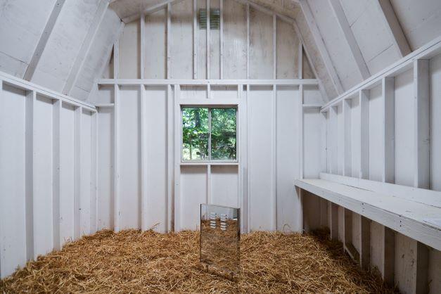 Interior of a small, barn-shaped building painted white with a window in the center and hay on the floor.