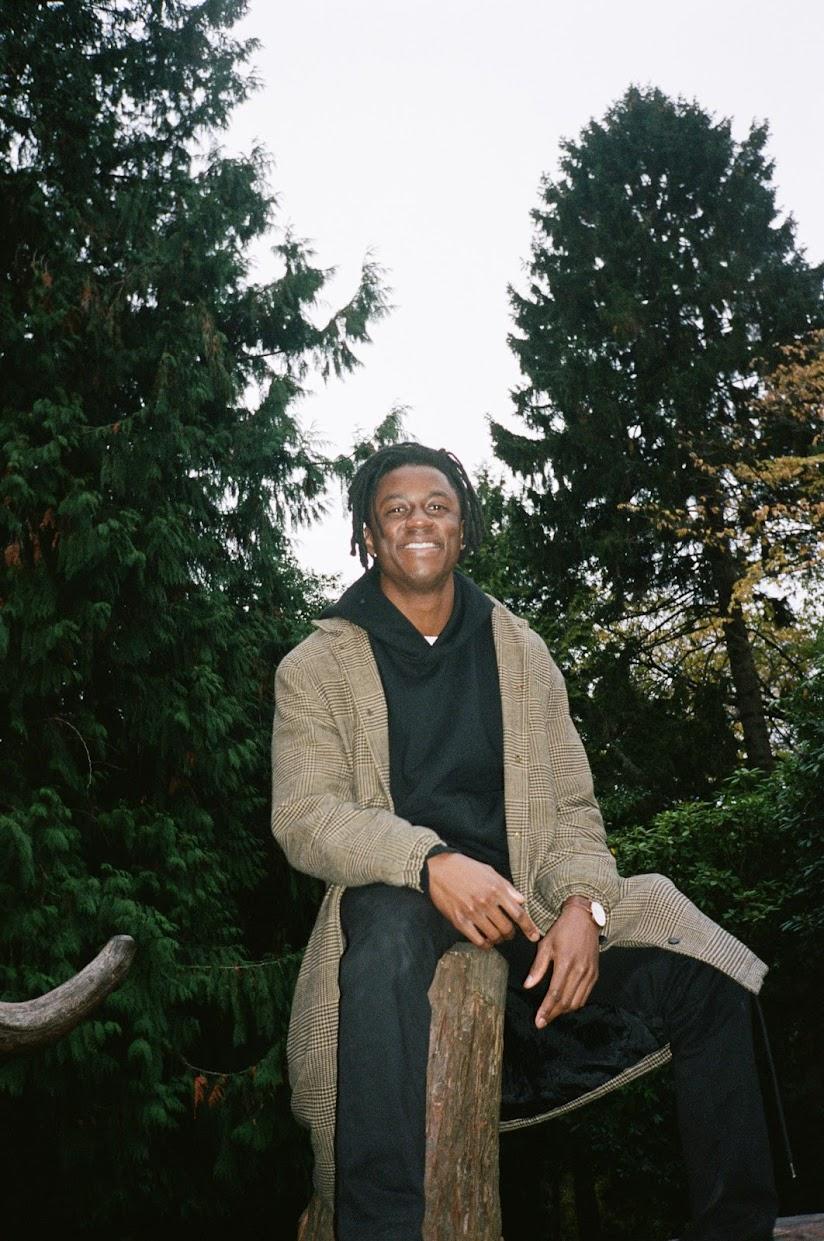 Photo of Jamal Yearwood, a young man with short black hair wearing a long tan coat, sitting on a wooden post among large evergreen trees
