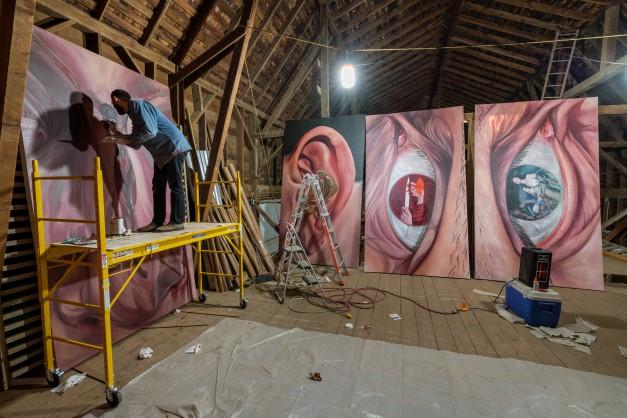 Man standing on scaffolding, painting a large-scale painting within a barn