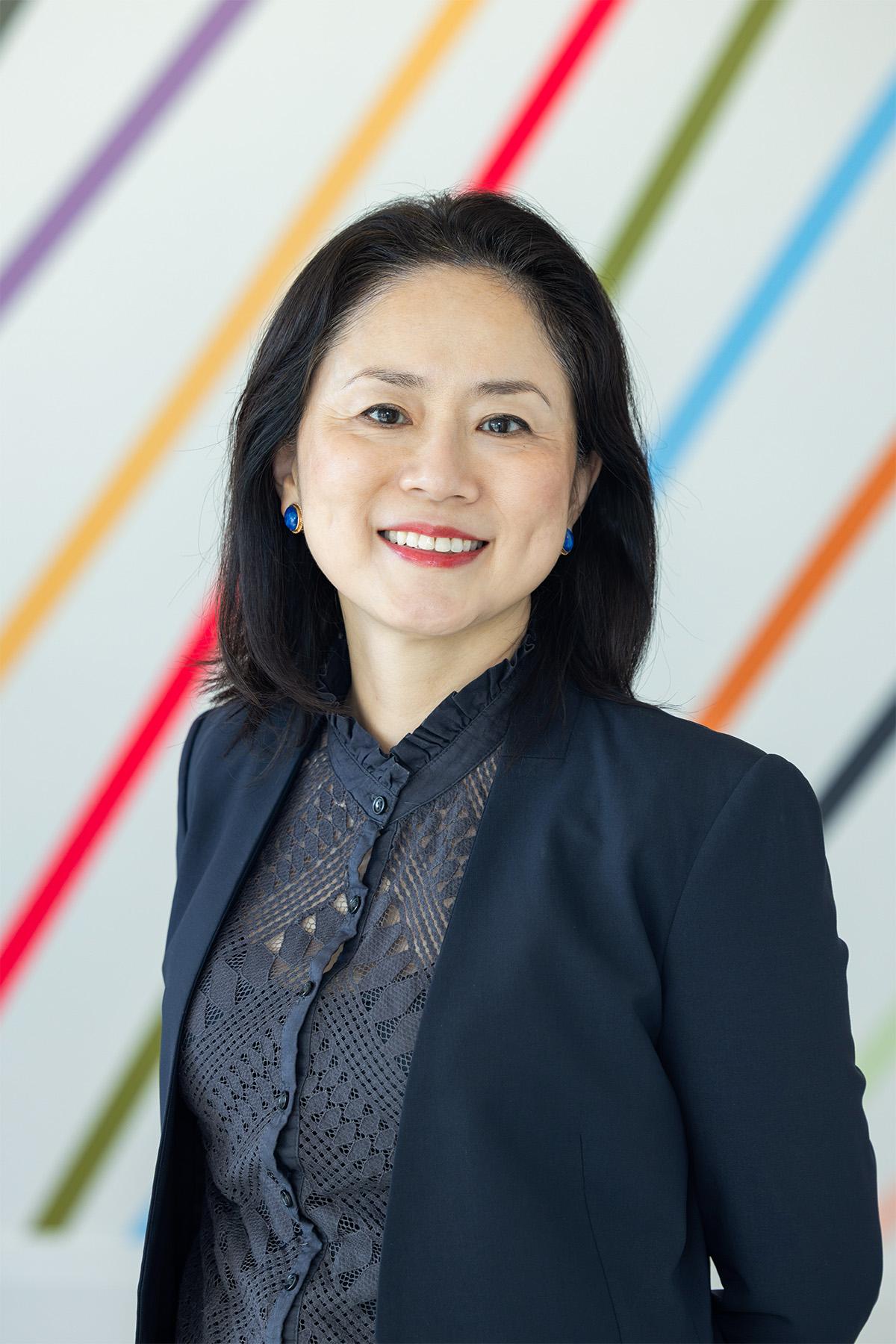 A professional headshot style photograph of a woman wearing formal clothing in front of a wall with colorful, diagonal stripes