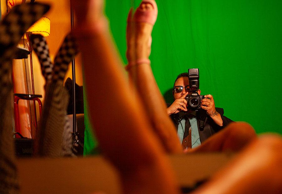 A person leaning against a green backdrop takes a photo with a large camera. In the foreground are mannequin legs